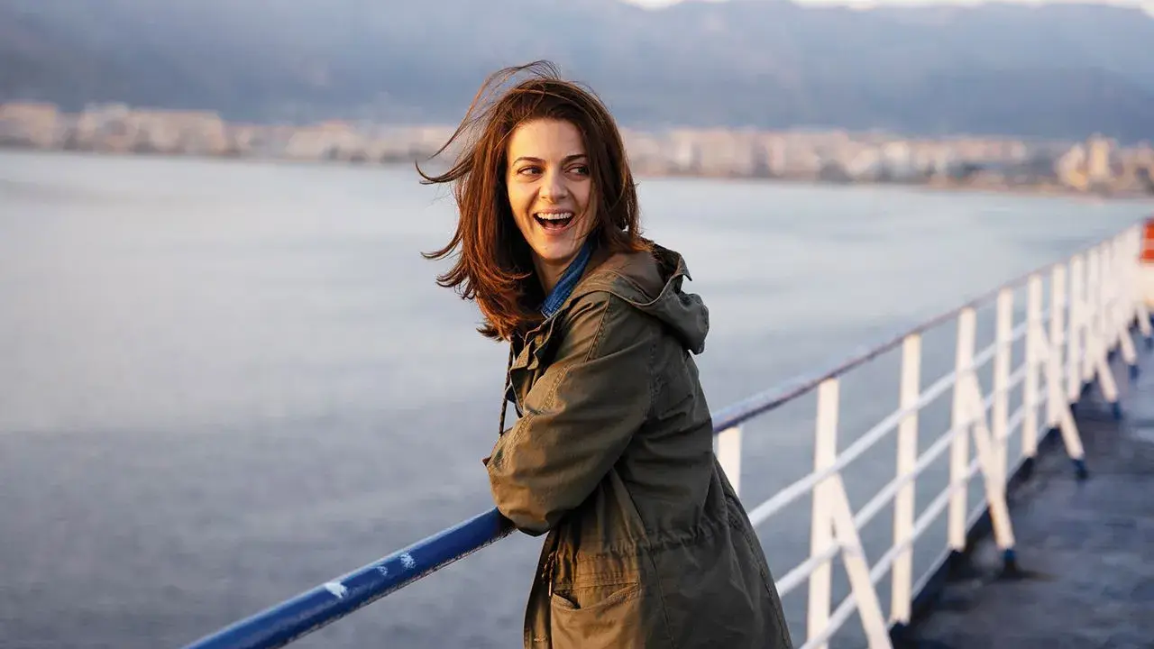 Una donna ride felice sul ponte di una nave, con la costa sullo sfondo. Sembra una scena da un **dieci minuti film** di avventura.