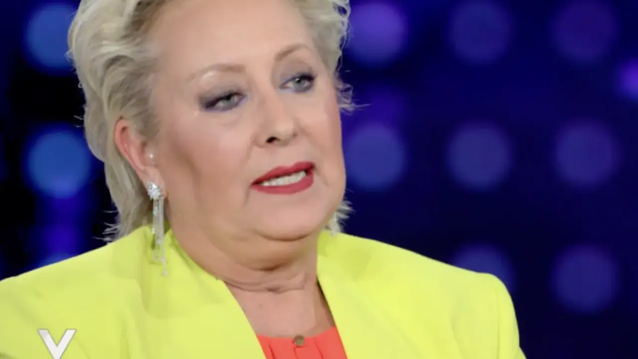 Carolyn Smith, come sta, con un tailleur giallo acceso e un top arancione, parla con un microfono.