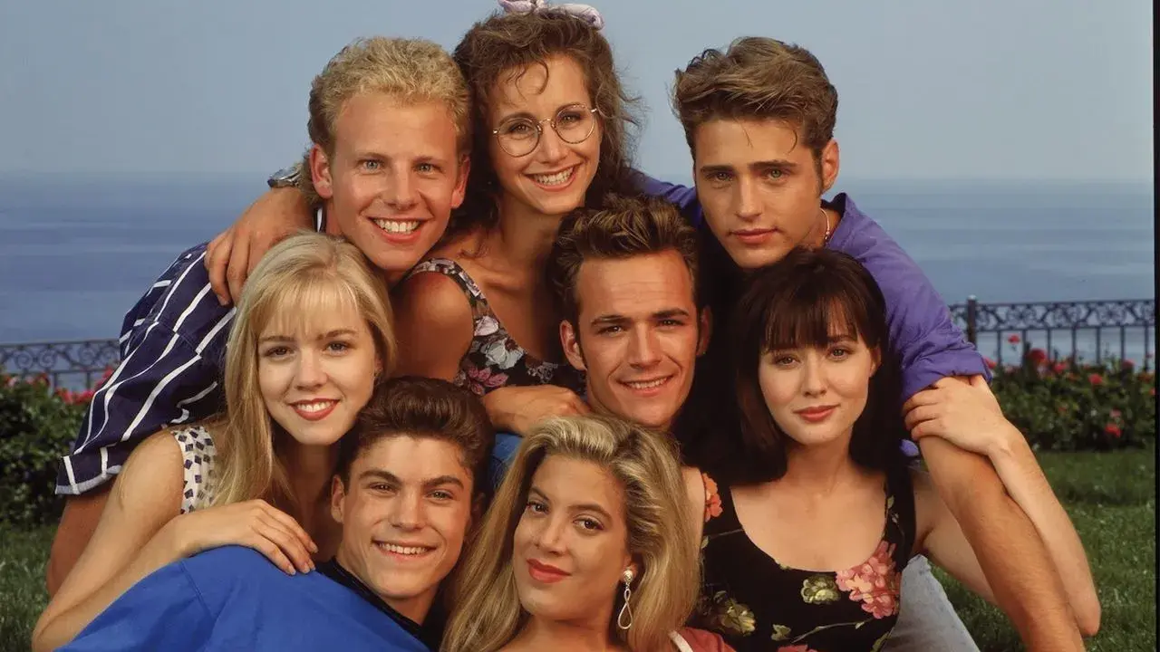 Il cast di Beverly Hills 90210, un gruppo di amici sorridenti con lo sfondo del mare.