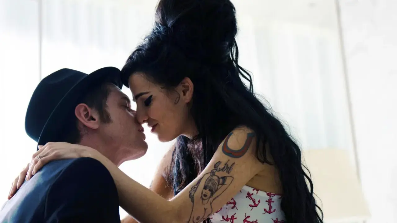 Blake Fielder-Civil, marito di Amy Winehouse, la bacia teneramente. Lei ha i capelli neri raccolti e indossa un top con ancore.