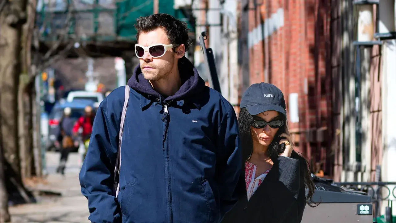 Harry Styles e la sua fidanzata passeggiano per strada, lui con occhiali bianchi, lei al telefono con un cappellino "KISS".