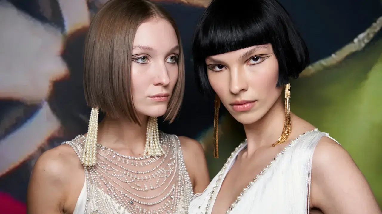 Due modelle con un sbarazzino caschetto corto sfilato, una con capelli castani e l'altra con capelli neri, sfoggiano orecchini pendenti e abiti decorati.