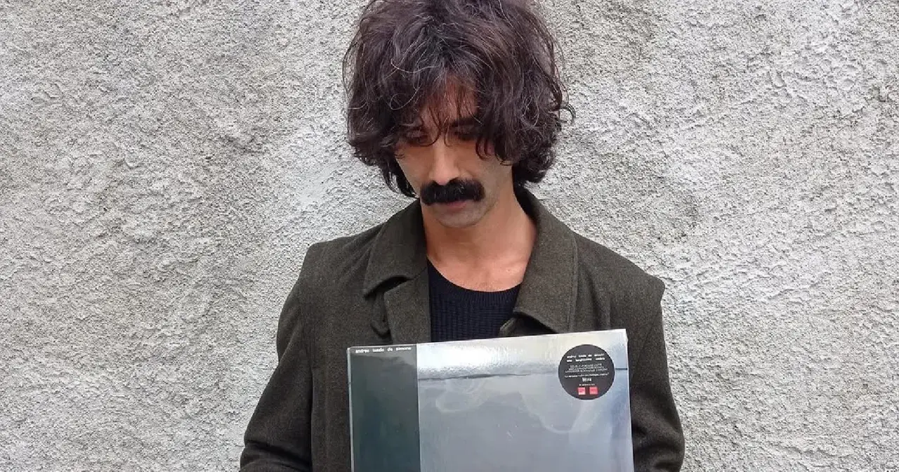 Andrea Laszlo De Simone con un disco in mano, con il suo iconico baffo e capelli ricci, davanti a un muro grezzo.