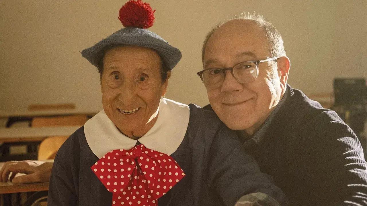 Un uomo con un cappello blu e un fiocco a pois rossi, sorride accanto a un uomo con gli occhiali. Un momento di gioia nella vita da Carlo.