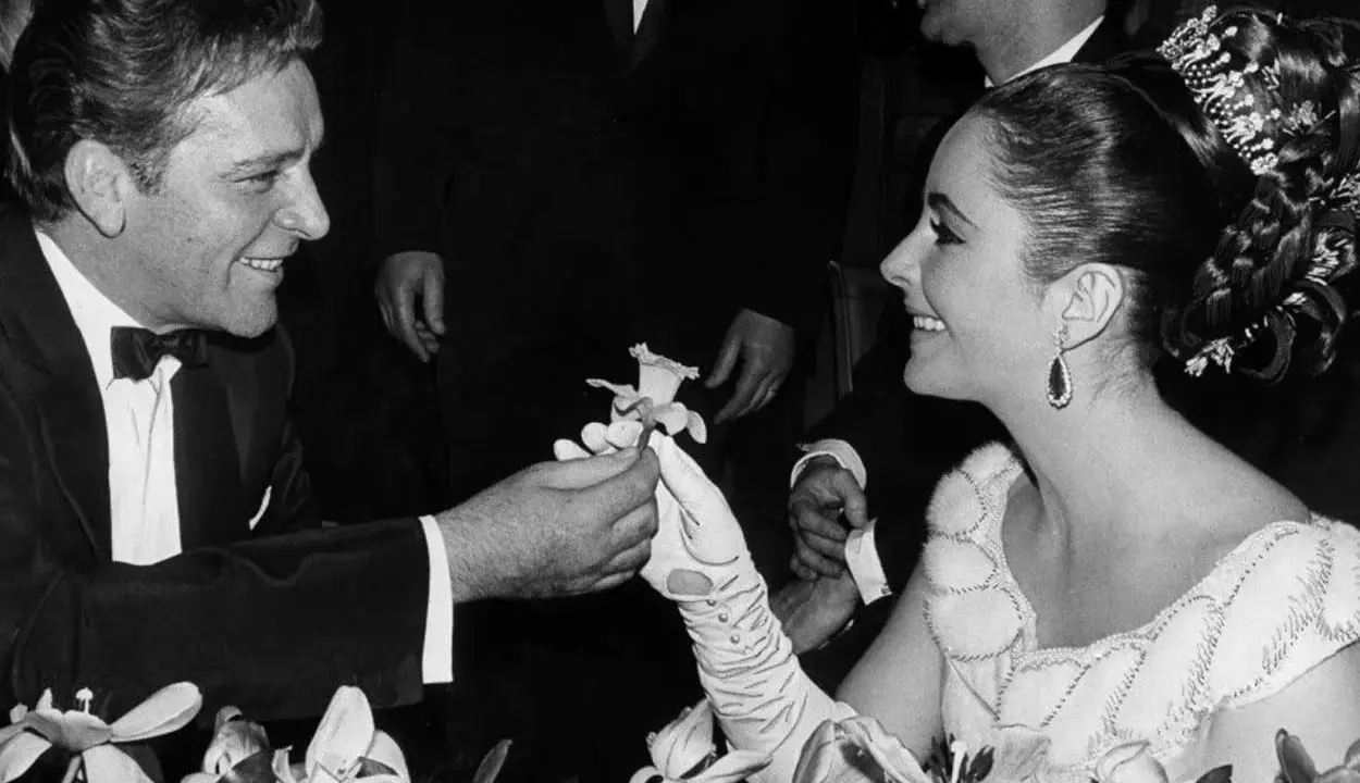 Richard Burton offre un fiore a Elizabeth Taylor, suo coniuge, in un momento di tenerezza tra fiori.