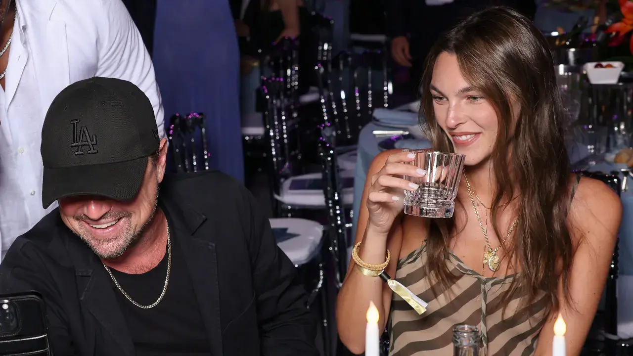 Vittoria Ceretti e Leonardo DiCaprio sorridono a una cena. Lei tiene un bicchiere, lui indossa un cappellino con la "LA".