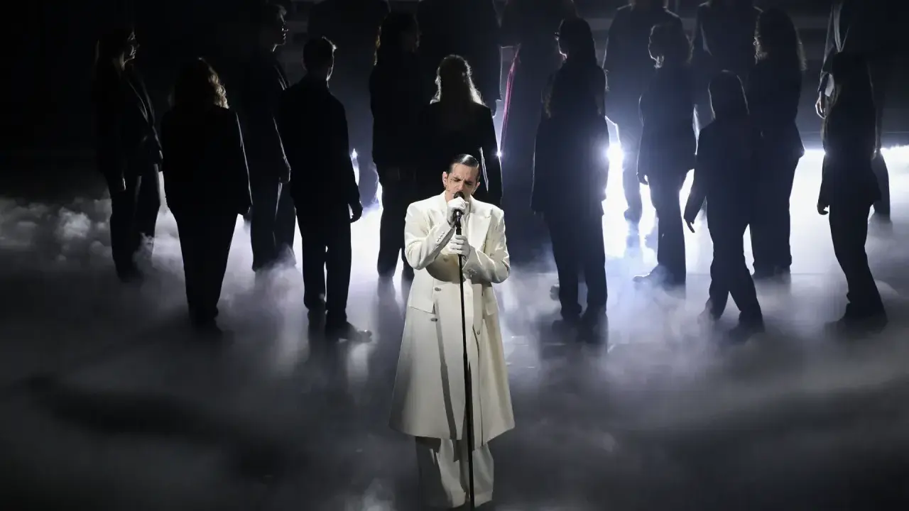Achille Lauro, vestito di bianco, canta sul palco circondato da un coro in controluce, un'immagine che evoca un sentimento di essere perdutamente innamorato.