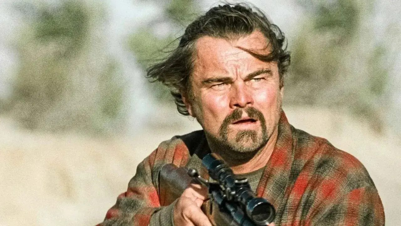Leonardo DiCaprio con fucile, in un film di sopravvivenza. Capelli mossi dal vento, sguardo intenso, camicia a quadri.