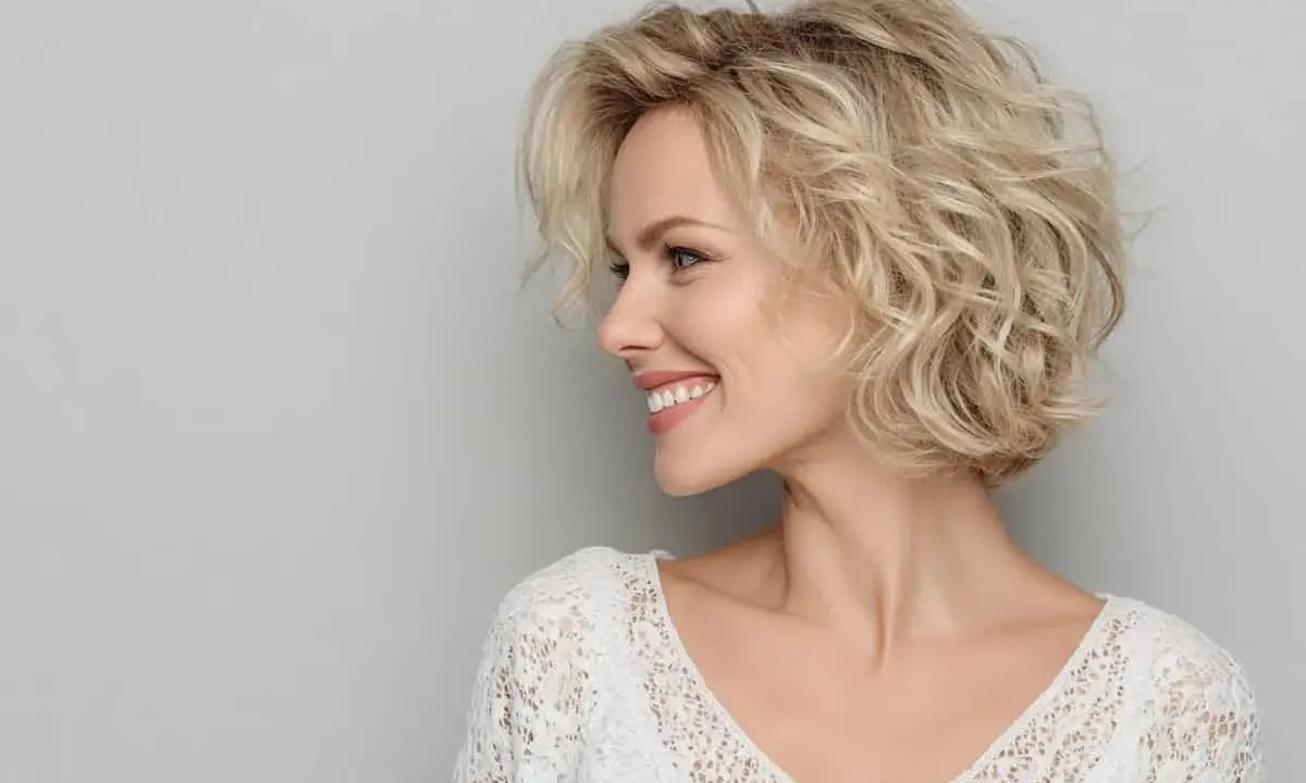 Donna sorridente con capelli biondi mossi, taglio **dietro sfumato caschetto corto**, indossa un top bianco in pizzo.