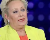 Carolyn Smith, come sta, con un tailleur giallo acceso e un top arancione, parla con un microfono.