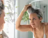 Donna si sistema i capelli grigi davanti allo specchio, cercando modi per camuffare i capelli bianchi senza tinta.