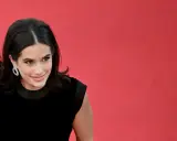 La figlia di John Travolta, Ella Bleu, sorride con un abito nero e orecchini scintillanti su sfondo rosso.