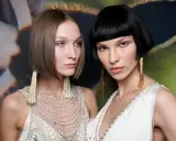 Due modelle con un sbarazzino caschetto corto sfilato, una con capelli castani e l'altra con capelli neri, sfoggiano orecchini pendenti e abiti decorati.