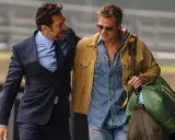 Brad Pitt 2024, con giacca gialla e camicia di jeans, cammina con un amico in completo scuro.
