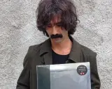 Andrea Laszlo De Simone con un disco in mano, con il suo iconico baffo e capelli ricci, davanti a un muro grezzo.