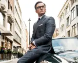 Taron Egerton, parte del cast di Kingsman 2, posa con eleganza su un'auto d'epoca, vestito in un impeccabile abito grigio.