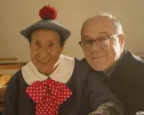 Un uomo con un cappello blu e un fiocco a pois rossi, sorride accanto a un uomo con gli occhiali. Un momento di gioia nella vita da Carlo.
