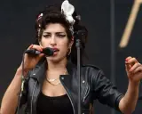 Amy Winehouse magra, con la sua iconica acconciatura e trucco, canta al microfono con passione. Indossa una giacca di pelle nera.