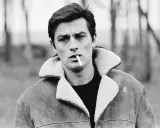 Alain Delon, iconico fisico, fuma una sigaretta con sguardo intenso, indossa una giacca di pelle con colletto di pelliccia.
