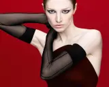 Hunter Schafer, prima donna con guanti neri velati e trucco drammatico, posa con le braccia incrociate su sfondo rosso.