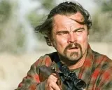 Leonardo DiCaprio con fucile, in un film di sopravvivenza. Capelli mossi dal vento, sguardo intenso, camicia a quadri.
