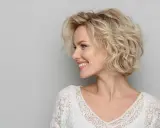 Donna sorridente con capelli biondi mossi, taglio **dietro sfumato caschetto corto**, indossa un top bianco in pizzo.