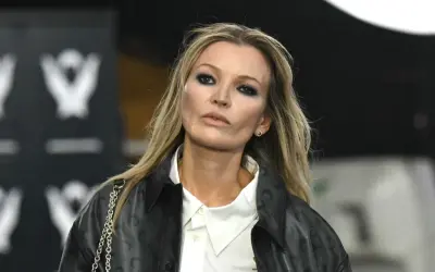 Kate Moss oggi, con un look audace e un trucco marcato, sfila con un giubbotto di pelle e una camicia bianca.