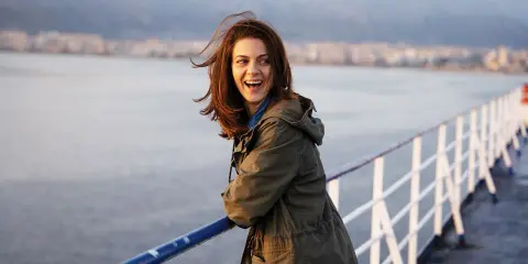 Una donna ride felice sul ponte di una nave, con la costa sullo sfondo. Sembra una scena da un **dieci minuti film** di avventura.
