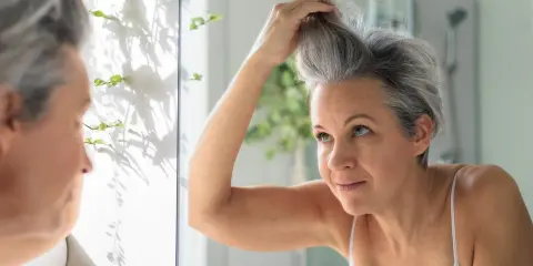 Donna si sistema i capelli grigi davanti allo specchio, cercando modi per camuffare i capelli bianchi senza tinta.