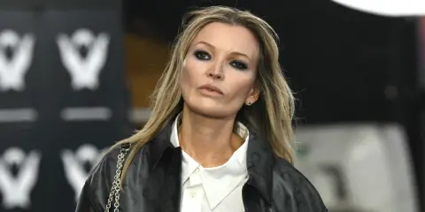 Kate Moss oggi, con un look audace e un trucco marcato, sfila con un giubbotto di pelle e una camicia bianca.