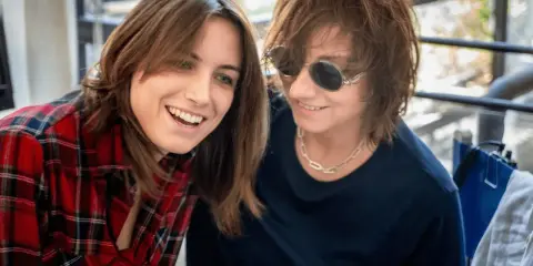 Due donne sorridono, una con camicia a quadri rossi, l'altra con occhiali da sole. Potrebbero essere Gianna Nannini e la sua moglie.