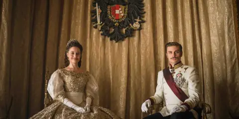 La principessa Sissi e l'imperatore Francesco Giuseppe nella serie tv, seduti su poltrone dorate, con uno stemma imperiale sullo sfondo.