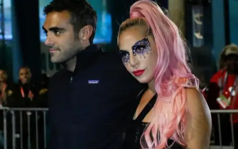 Lady Gaga con il suo fidanzato, Christian Carino, durante un evento. Lei indossa un trucco scintillante e capelli rosa.