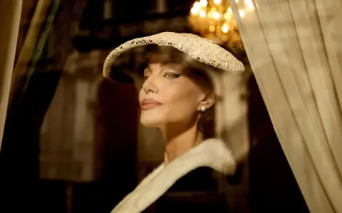 Angelina Jolie nei panni di Maria Callas, con un cappello di pizzo e una pelliccia, evoca l'eleganza di un'epoca passata.