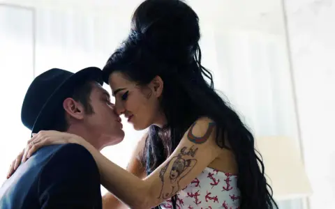 Blake Fielder-Civil, marito di Amy Winehouse, la bacia teneramente. Lei ha i capelli neri raccolti e indossa un top con ancore.