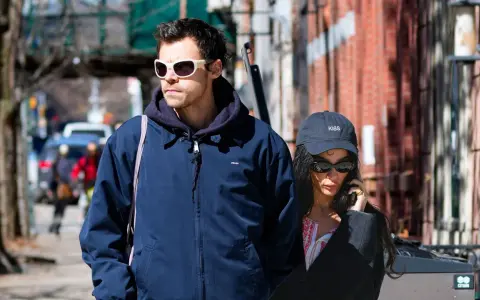 Harry Styles e la sua fidanzata passeggiano per strada, lui con occhiali bianchi, lei al telefono con un cappellino "KISS".