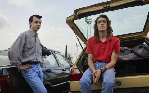 Due ragazzi in jeans e camicie, appoggiati a due auto d'epoca. Scena dalla serie tv 883.