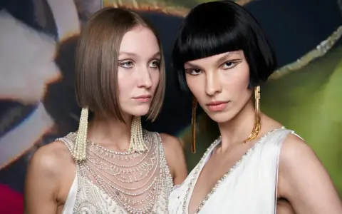 Due modelle con un sbarazzino caschetto corto sfilato, una con capelli castani e l'altra con capelli neri, sfoggiano orecchini pendenti e abiti decorati.