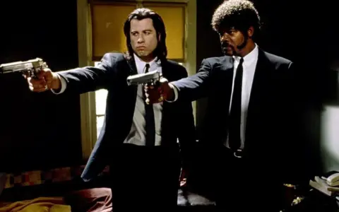 Due uomini in abito scuro puntano pistole, iconica scena da un film anni 90.