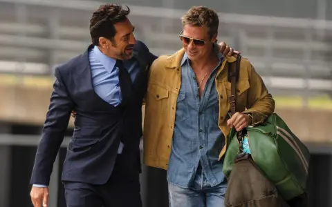 Brad Pitt 2024, con giacca gialla e camicia di jeans, cammina con un amico in completo scuro.
