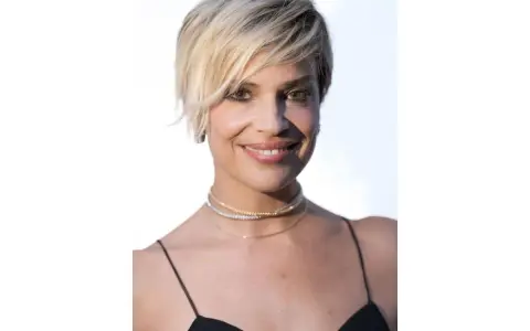 Donna con capelli cortissimi biondi, look 2026, indossa un abito nero e collane scintillanti.