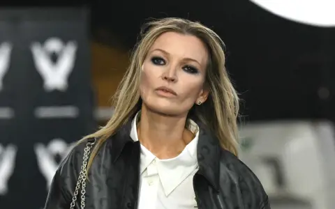 Kate Moss oggi, con un look audace e un trucco marcato, sfila con un giubbotto di pelle e una camicia bianca.