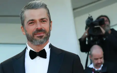 Uomo con capelli bianchi e barba, indossa un abito elegante con papillon nero. Dietro di lui, un operatore riprende con una telecamera, quasi a voler dimostrare che i capelli bianchi possono tornare neri.