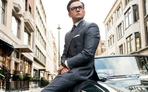 Taron Egerton, parte del cast di Kingsman 2, posa con eleganza su un'auto d'epoca, vestito in un impeccabile abito grigio.
