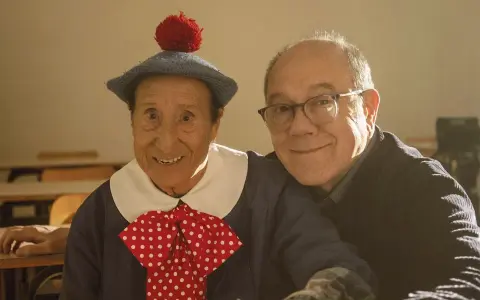 Un uomo con un cappello blu e un fiocco a pois rossi, sorride accanto a un uomo con gli occhiali. Un momento di gioia nella vita da Carlo.