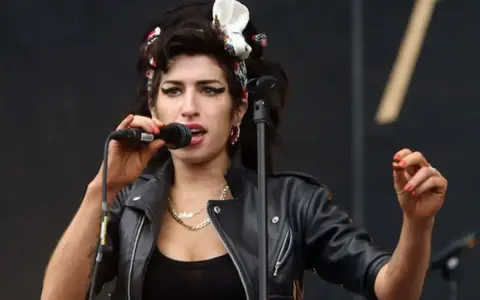 Amy Winehouse magra, con la sua iconica acconciatura e trucco, canta al microfono con passione. Indossa una giacca di pelle nera.