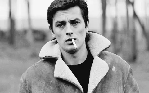 Alain Delon, iconico fisico, fuma una sigaretta con sguardo intenso, indossa una giacca di pelle con colletto di pelliccia.