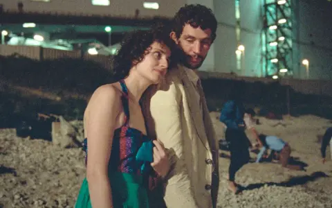 Il cast de "La Chimera" in un momento intimo, con altre figure sfocate sullo sfondo notturno.