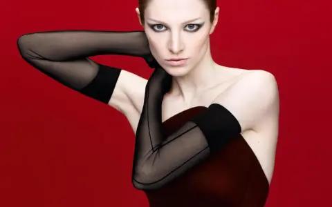 Hunter Schafer, prima donna con guanti neri velati e trucco drammatico, posa con le braccia incrociate su sfondo rosso.