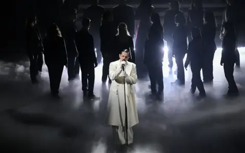 Achille Lauro, vestito di bianco, canta sul palco circondato da un coro in controluce, un'immagine che evoca un sentimento di essere perdutamente innamorato.